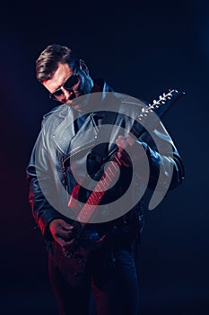 Brutal bearded Heavy metal musician in leather jacket, cap and sunglasses is playing electrical guitar. Shot in a studio