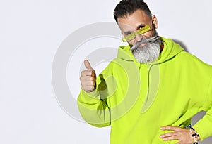 Brutal bearded middle aged hipster man in bright yellow green hoodie and glasses looking at camera and showing thumb up sign