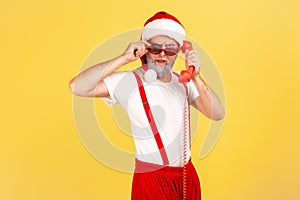 Brutal grey bearded adult man in santa claus hat holding handset of landline telephone and looking at camera through sunglasses,