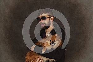 Brutal guy grins in sunglasses, with a beard and fashionable hairstyle and holding a dog