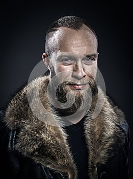 Brutal handsome smiling unshaven man with long beard and moustache in black fur coat with collar.