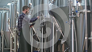 Brutal Male brewer with a beard controls the readings of devices on beer tanks using a tablet. Craft beer production