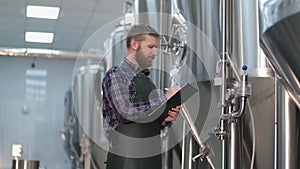 Brutal a male brewer with a beard stands near beer tanks and records instrument readings. Craft beer production