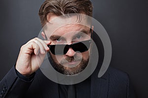 A brutal man with a beard and a stylish hairstyle in a black suit looks appallingly at the camera, dropping his sunglasses