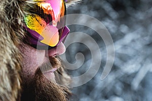 A brutal man with a beard in a winter hat and ski goggles looks into the distance while standing in the mountains in