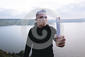 Brutal man in black windbreaker holding blue flare torch in hand, standing on top of rock in evening with view on river.  Ultras