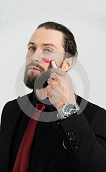 A brutal man paints red lipstick on his cheek. Dressed in a suit and red tie.