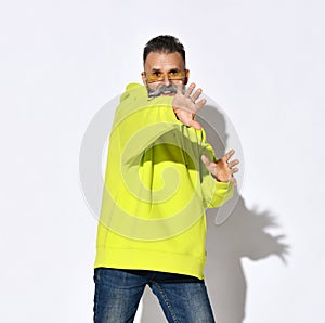 Brutal smiling bearded middle aged man in yellow hoodie and jeans standing and gesticulating with hands on camera