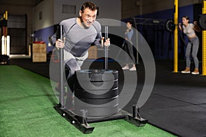 Brutal strong muscular bodybuilder athletic man pumping up muscles with crossfit traction sled in gym. Workout