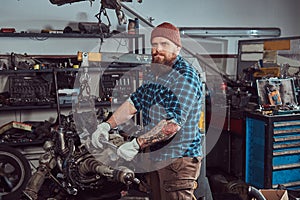 A brutal tattooed bearded mechanic specialist repairs the car engine which is raised on the hydraulic lift in the garage