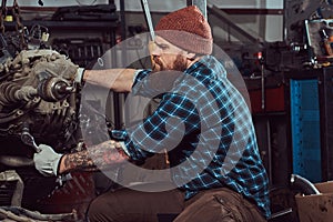 A brutal tattooed bearded mechanic specialist repairs the car engine which is raised on the hydraulic lift in the garage