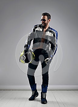 Brutal unshaved adult man biker motocross racer in motorcycle gear boots, jacket and gloves walks with helmet in hand
