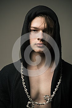 Brutal young man wearing hooded black outfit and bicycle around his neck isolated on gray background, troublemaker and photo