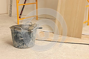 Bucket of adhesive mix with spatula near tile on floor indoors