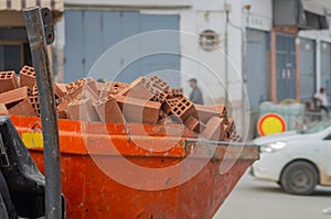 The bucket of a mini loader, which carries a fully loaded red brick down the street. An example of solving problems with low costs