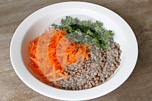Buckwheat porridge with grated carrots and parsley in white plate on wooden table background
