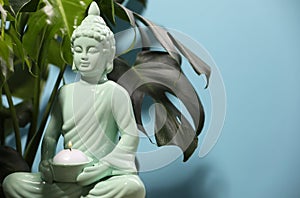 Buddhism religion. Decorative Buddha statue with burning candle and monstera against light blue wall, space for text
