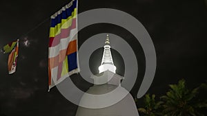 Buddhist stupa stands tall in moonlight, fluttering prayer flags, sacred site illuminated at night. Palm trees sway