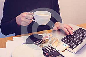 Budget planing concept. Side view of a woman drinking coffee while summing up her budget with the help of calculator and a laptop