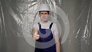 Builder in blue overalls puts on white hard hat and shows thumbs up.