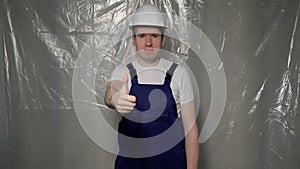 Builder in blue overalls puts on white hard hat and shows thumbs up.