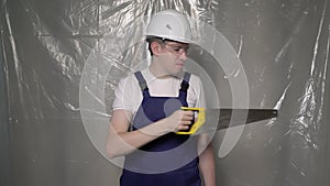 Builder in blue overalls and white hard hat helmet and glasses with hand saw.