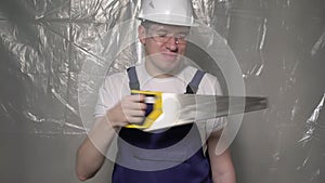 Builder in blue overalls and white hard hat helmet and glasses with hand saw.