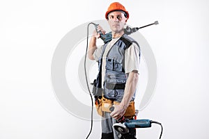 A builder with a hammer drill on his shoulder, and a angle grinder in his other hand, in a helmet, smiles