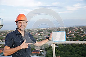 Builder in a protective helmet with a tablet for writing in his hand is standing on the roof of a building overlooking the city