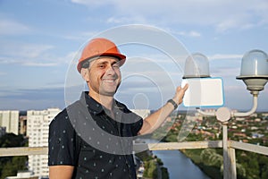 Builder in a protective helmet with a tablet for writing in his hand is standing on the roof of a building overlooking the city