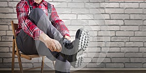 Builder putting on shoes for work near brick wall.