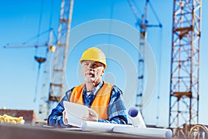 Builder in reflective vest and hardhat standing at construction site and holding