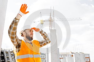 Builder repairman, foreman in safety helmet and vest standing at his workplace in a building under construction and and making a