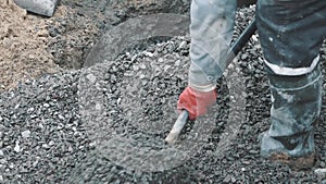 Builder in rubber boots rake macadam with shovel in sewer ditch at building site
