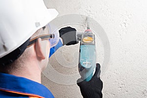 Builder in safety glasses and helmet drills a wall with a hammer drill, rear view