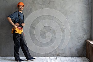 A builder stands near a gray wall wearing an orange hard hat and a tool belt. Concrete wall and constructor. Repairman