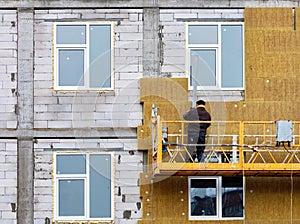 The builder stands on a suspended cradle and insulates the facade of the house under construction with mineral insulation