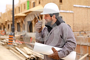 Builder talking by walkie talkie and holding drawing plan at construction site.