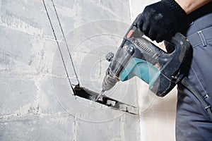 Builder worker man with hammer drill perforator equipment making hole in gray wall construction