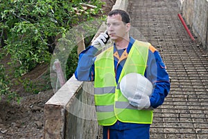 Builder in a yellow vest discusses by phone the construction time standing on the background of the stairs