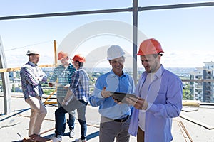 Builders On Site Holding Tablet Computer Discuss Construction Project Over Team Of Apprentices Talking