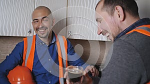 Builders in working clothes and protective helmets, drinking coffee in the kitchen during a break.