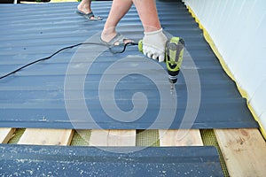 A building constructor is installing lightweight metal roofing sheet using an electric screw driver on the roof covered with photo