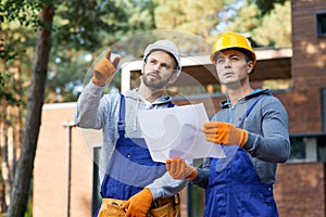 Building ideas. Portrait of two builders looking away while standing outdoors with an open blueprint discussing the half