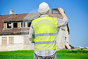 Building inspector filming on tablet PC near old abandoned, damaged house on grass field