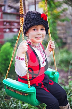 Bulgarian boy in ethnic folklore costume with traditional embroidery on a swing in a garden at his family house