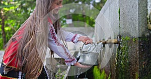 Bulgarian girl in traditional folklore attire, ethnic slavic clothing pouring water from old stone tap or fountain in Bulgaria