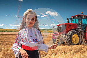 Bulgarian Girl in traditional folklore costume in agricultural wheat field during harvest time with industrial tractor machine
