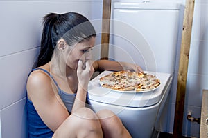 Bulimic woman feeling sick guilty sitting at the floor of the toilet leaning on WC eating pizza