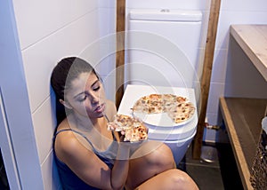 Bulimic woman feeling sick guilty sitting at the floor of the toilet leaning on WC eating pizza